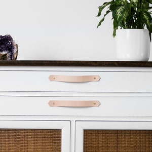 Puede incluir: Cómoda blanca pintada con dos cajones y dos puertas de armario. Las puertas del armario tienen un panel de ratán marrón tejido. La cómoda tiene tiradores de cuero con detalles en latón.