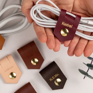 May include: Six leather cord organizers with gold accents and personalized names: Kestrel, Addison, Travis, Stephanie, N.E.Mc., and a burgundy organizer with a white cord.