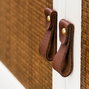 May include: Close-up of two brown leather cabinet pulls with gold accents against a white and brown woven background.