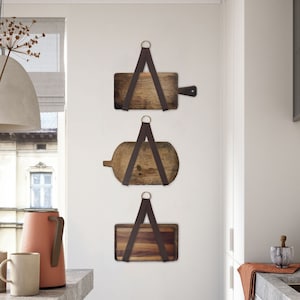 Puede incluir: Tres tablas de cortar de madera con correas de cuero cuelgan de una pared blanca en una cocina. Las tablas son de diferentes tamaños y formas. La cocina tiene gabinetes blancos y una encimera gris.