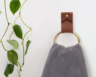 May include: A grey towel hanging on a gold ring attached to a brown leather hook on a white wall. A green plant is hanging on the left side of the wall.