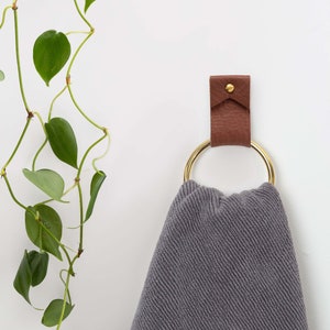 May include: A gray towel hanging on a gold ring attached to a brown leather hook on a white wall. A green plant is hanging on the left side of the wall.