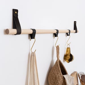 Op de afbeelding: Een houten keukenrail met zwarte leren banden en goudkleurige haken. Een beige mesh tas, een geweven mand en maatlepels hangen aan de haken. De rail is aan een witte muur bevestigd, wat wijst op een keukenorganisatiethema.
