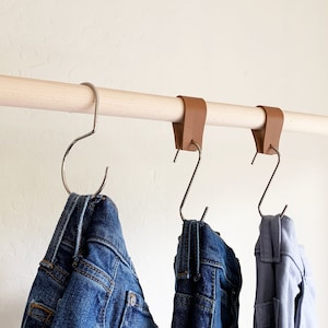 Puede incluir: Tres pares de pantalones colgados de una barra de madera con correas de cuero. Los pantalones son de mezclilla azul y gris.