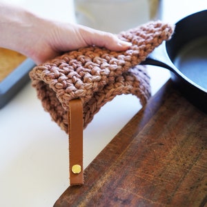 May include: A brown crocheted pot holder with a leather strap and a gold button. The pot holder is being used to lift a cast iron skillet.