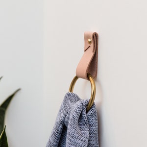 May include: A leather and brass wall hook with a light tan leather strap and a large brass ring. A gray and white patterned towel is hanging from the ring. The hook is mounted on a white wall.