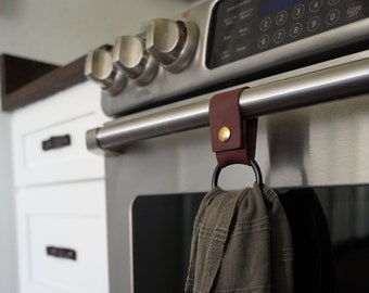 May include: A brown leather towel holder with a gold button is attached to a stainless steel oven handle. A dark green towel hangs from the black ring. The kitchen appliance has a digital display and white cabinets.