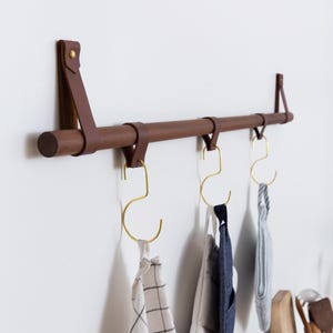 Peut inclure: Un porte-serviettes mural en bois avec des sangles en cuir marron et trois crochets dorés. Le porte-serviettes a trois torchons accrochés, un blanc à carreaux noirs, un bleu à rayures blanches et un blanc à rayures bleues.