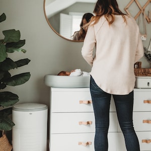 May include: A white chest of drawers with three drawers and brown leather handles. A baby is lying on a grey changing mat on top of the dresser. A woman is standing behind the dresser, looking at the baby. The dresser is in a nursery with a white nappy bin and a green plant in a wicker basket.