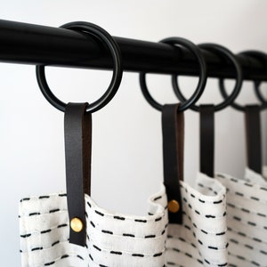 May include: Black metal curtain rod with black rings and brown leather tabs. The curtains are white with black stitching.