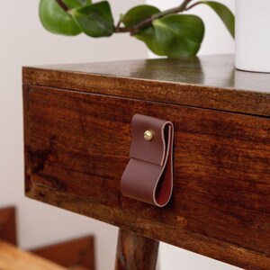 May include: A close-up of a wooden drawer with a dark brown leather pull. The drawer is part of a wooden table with a plant in a white pot on top. The leather pull is secured with a gold-colored rivet.