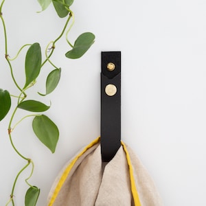 May include: A black leather towel hook with brass hardware, holding a beige towel with a yellow trim. A green leafy plant is in the background, creating a minimalist aesthetic. The hook is mounted on a white wall.