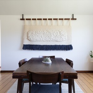 May include: A white and blue woven wall hanging with a fringe border hangs above a dark wood dining table with four chairs.