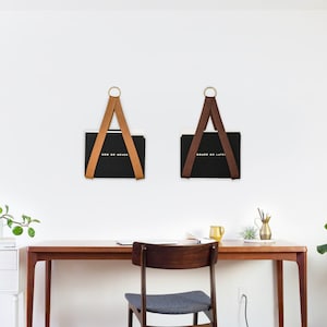 Può includere: Un ambiente di lavoro per ufficio a casa con una scrivania e una sedia in legno. Due cartelle nere sono appese al muro con cinturini in pelle e anelli in ottone. Le cartelle hanno il testo "Done or Never" e "Now or Never" stampato su di esse.