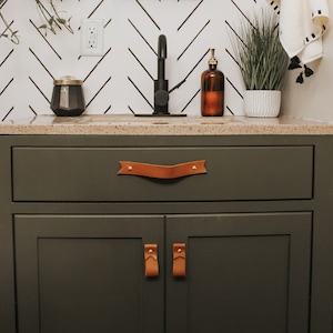 Könnte beinhalten: Ein dunkelgrüner Badezimmer-Waschtisch mit einem schwarzen Wasserhahn und einem braunen Leder-Schubladenzug. Der Waschtisch hat zwei Türen mit braunen Leder-Griffen. Der Waschtisch steht vor einer weißen und schwarzen Zickzack-Musterwand.