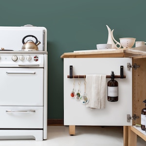 May include: A white kitchen with a vintage stove and a cabinet. The cabinet has a wooden towel rack, a brown bottle, and decorative ornaments. A beige towel hangs on the rack. The background is a dark teal color.