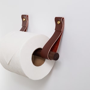 May include: A brown leather toilet paper holder with a wooden dowel and brass accents. The holder is mounted to a white wall.