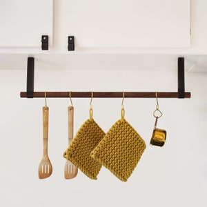 May include: A dark brown wooden kitchen utensil rack with four hooks. Two yellow knitted pot holders hang from two of the hooks. Two wooden spatulas hang from the other two hooks. A small gold metal measuring cup hangs from one of the hooks.