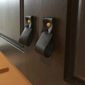 以下が含まれることがあります： 金色の金属製のアクセントが付いた黒い革製のキャビネットの取っ手が2つ。取っ手は濃い茶色のキャビネットのドアに取り付けられています。
