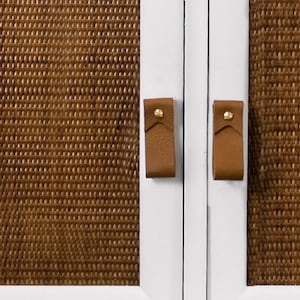 May include: White cabinet with two doors. Each door has a brown woven panel and a leather strap handle with a brass accent.