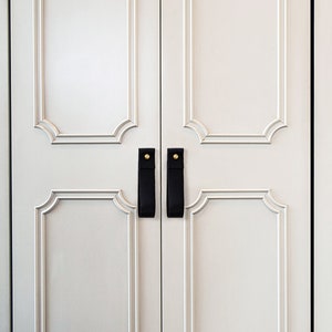 May include: White closet doors with ornate molding and black leather pulls with gold accents.