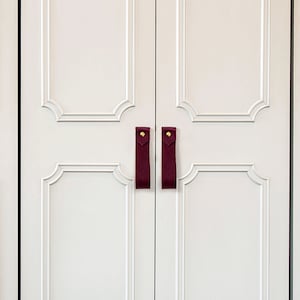 May include: Close-up of a white cabinet with decorative molding and burgundy leather strap handles. The cabinet doors are closed, and the handles are attached with gold-colored rivets. The overall design is clean and modern.