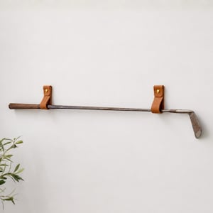 Op de afbeelding: Een vintage golfclub die horizontaal aan een muur is bevestigd met twee lichtbruine leren riemen. Elke riem is vastgezet met een gouden metalen klinknagel, waardoor de donkere houten steel en de metalen clubkop in een vaste positie hangen.