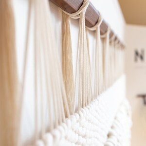 May include: A white macrame wall hanging with a wooden dowel at the top. The macrame is made with thick white yarn and has a textured, woven pattern.