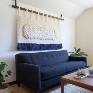 May include: A blue sofa with a white and blue woven wall hanging above it. A wooden coffee table with a plant and books on it sits in front of the sofa.