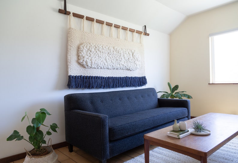 May include: A blue sofa with a white and blue woven wall hanging above it. A wooden coffee table with a plant and books on it sits in front of the sofa.