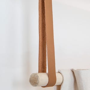 May include: A tan leather strap is looped over a wooden dowel, which is holding up a white fabric. The leather strap is attached to the ceiling. The dowel is made of light-colored wood.