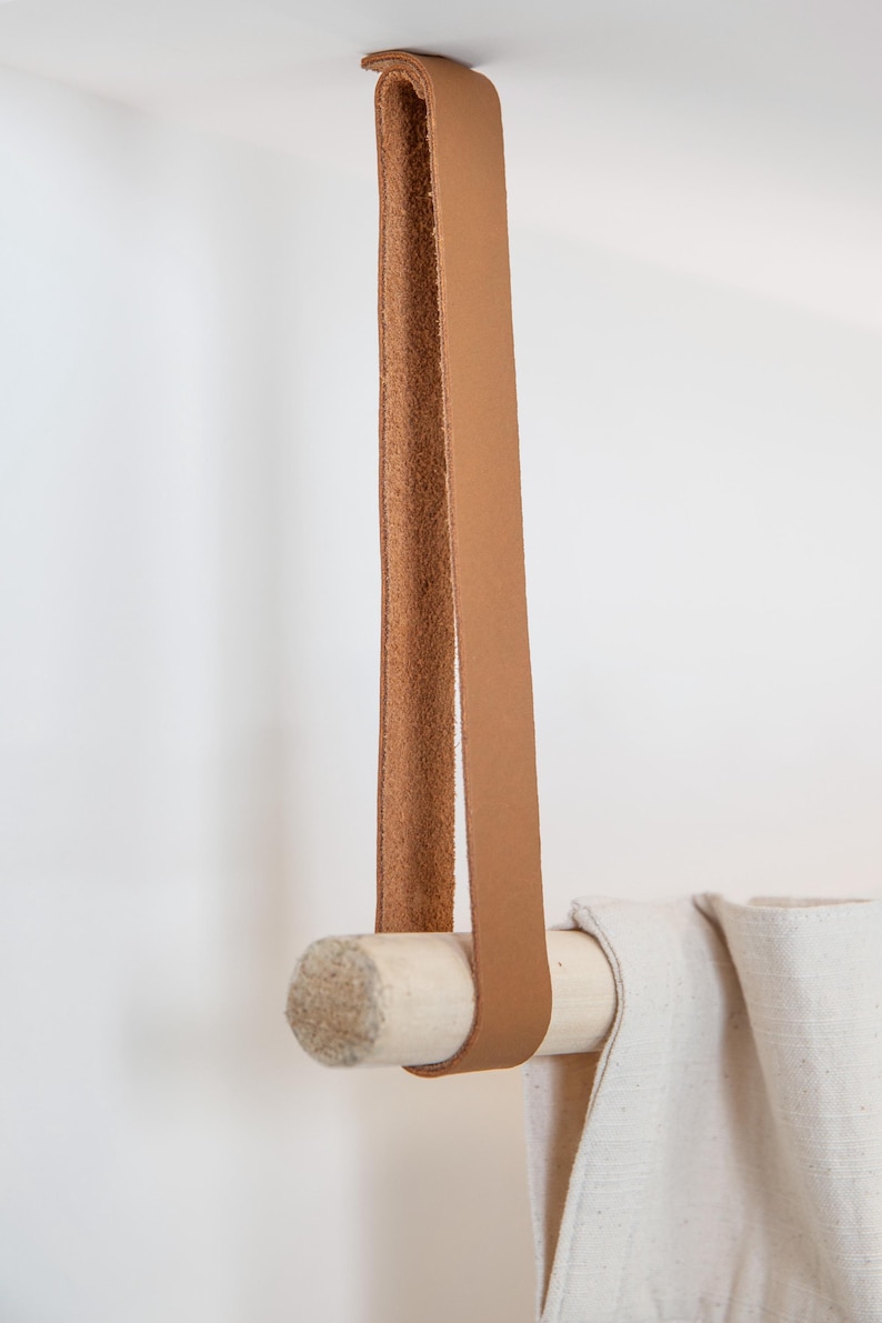 May include: A tan leather strap is looped over a wooden dowel, which is holding up a white fabric. The leather strap is attached to the ceiling. The dowel is made of light-colored wood.