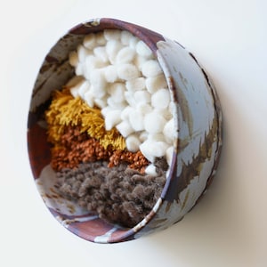 May include: A ceramic bowl with a textured, blue and brown glaze. The bowl is filled with a variety of colorful yarn and wool, including white, brown, orange, and yellow.