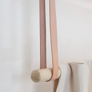 May include: A light-colored leather strap and wooden dowel curtain rod. The rod is suspended from the ceiling by a loop of light brown leather. A cream-colored curtain is draped over the rod.