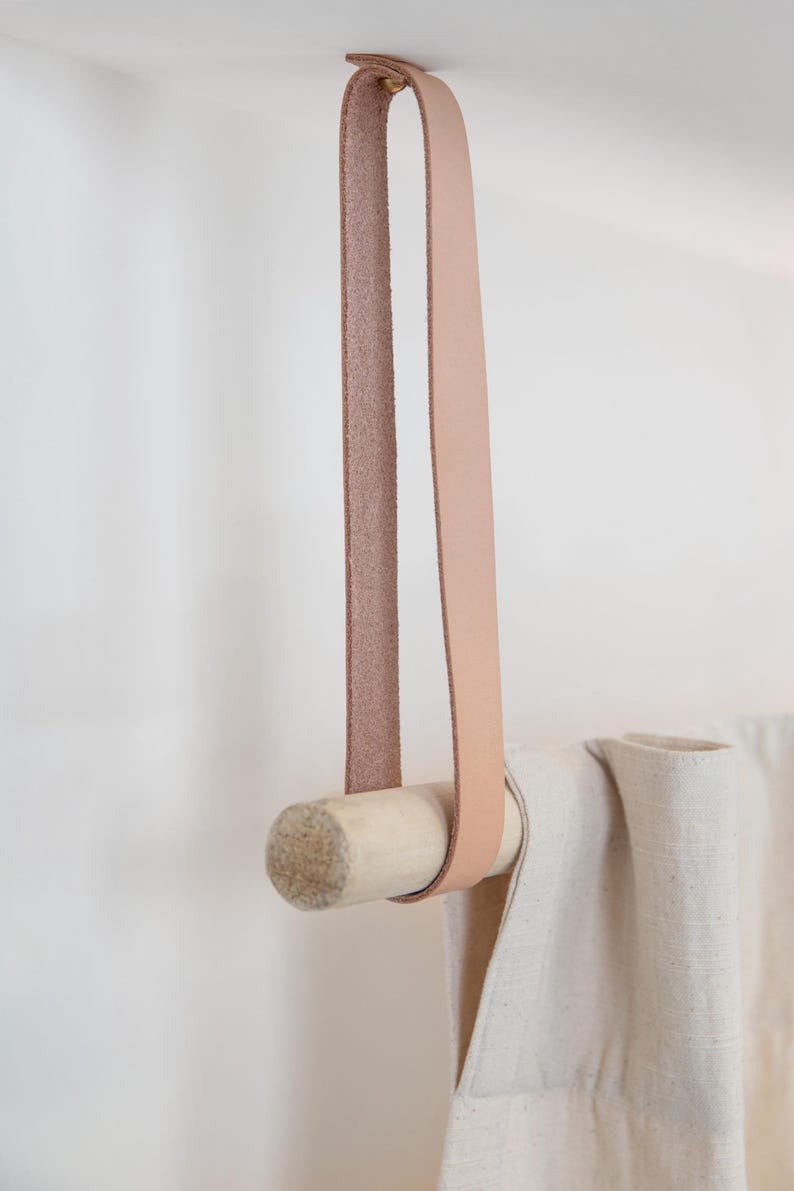 May include: A light-colored leather strap and wooden dowel curtain rod. The rod is suspended from the ceiling by a loop of light brown leather. A cream-colored curtain is draped over the rod.