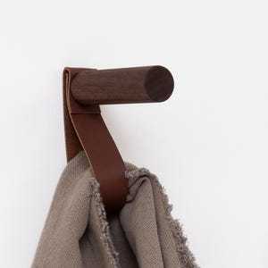 Op de afbeelding: Een aan de muur gemonteerde houten haak met een donkerbruine afwerking en een leren riem. Een grijze textiel hangt aan de haak. De haak is ontworpen om handdoeken, kleding of andere items op te hangen. Het ontwerp is minimalistisch en modern.