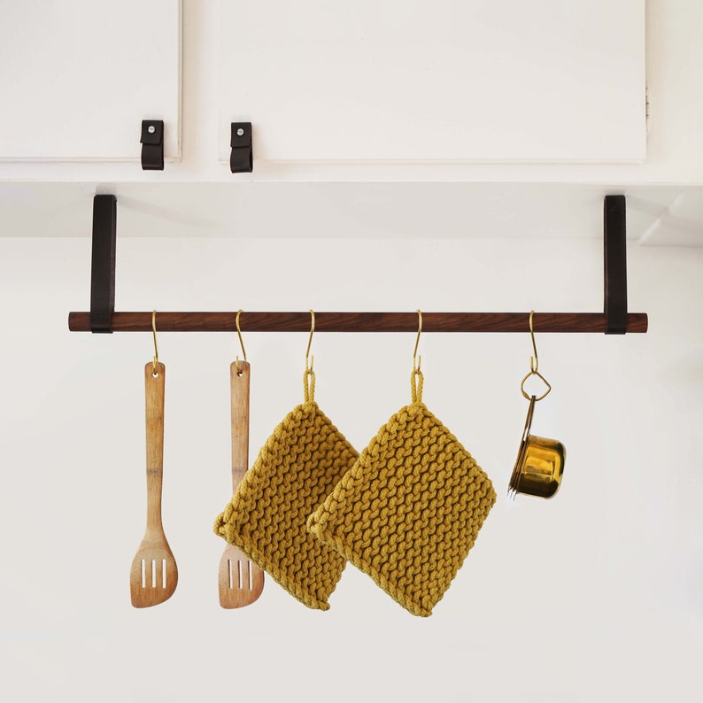 May include: A dark brown wooden kitchen utensil rack with four hooks. Two yellow knitted pot holders hang from two of the hooks. Two wooden spatulas hang from the other two hooks. A small gold metal measuring cup hangs from one of the hooks.