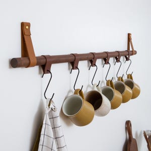 Könnte beinhalten: Ein Wand-Tassenhalter aus Holz mit Lederriemen und Haken. Sechs Keramikbecher hängen an den Haken, und ein kariertes Geschirrtuch hängt an einem der Haken.