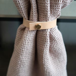 Neutral kitchen towel gathered with a natural veg-tan leather strap, handcrafted from vegetable-tanned full-grain leather using eco-friendly tanning processes. The brass screw closure and minimalist design reflect Keyaiira’s sustainable approach to modern home essentials.