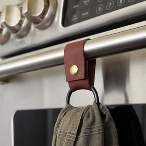 May include: A burgundy leather towel holder with a gold-colored snap is attached to a stainless steel oven handle. A black metal ring holds a brown and tan patterned kitchen towel.