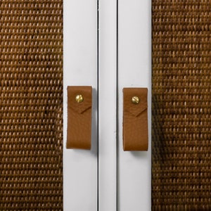 May include: Two brown leather cabinet pulls with gold screws on a white cabinet door.