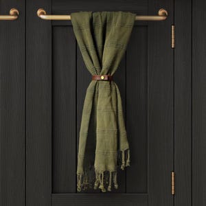 May include: A dark green striped towel with fringe hanging on a brass towel bar. The towel is secured with a brown leather strap with a brass buckle.