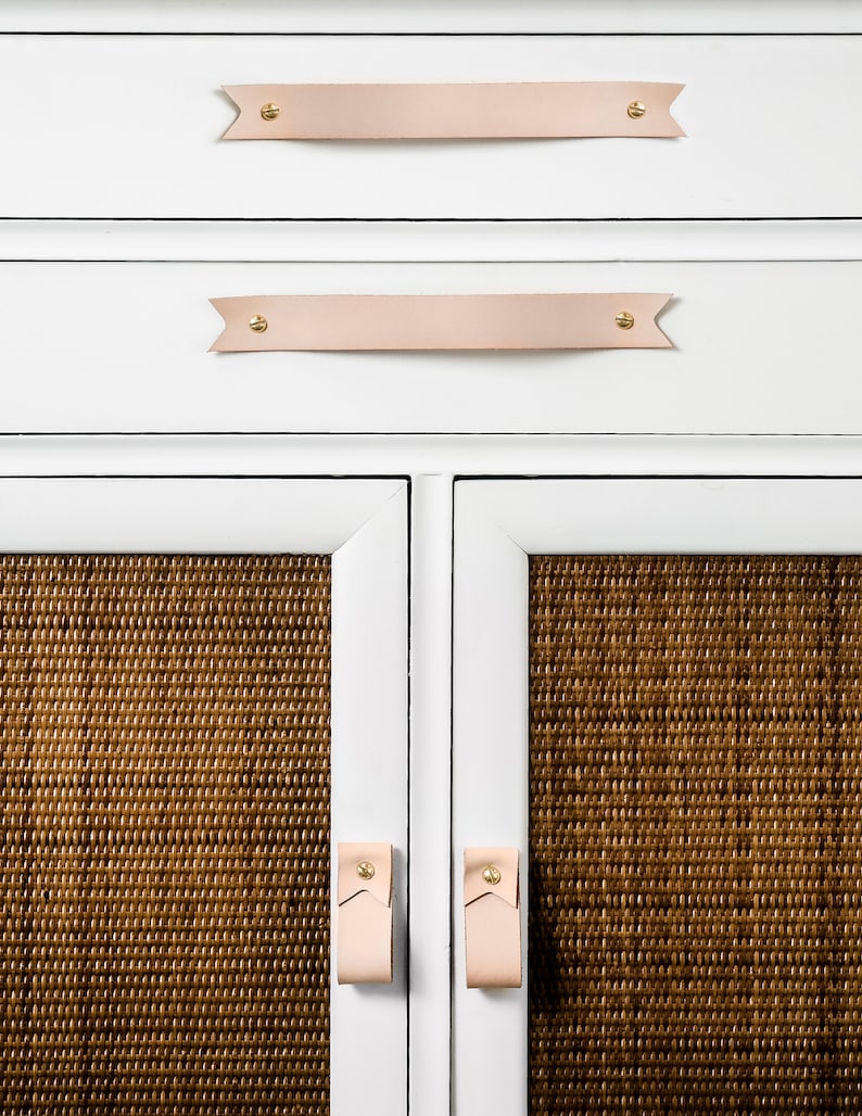 Keyaiira Veg Tan genuine leather drawer handle with V-shaped pointed ends and polished brass mounting hardware installed horizontally on a white cabinet above woven cane doors, highlighting the light natural leather color and handcrafted furniture design
