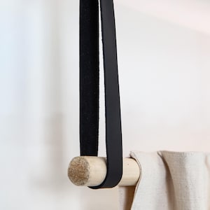May include: A black leather strap with a wooden dowel, designed for hanging a curtain or towel. The strap is attached to the ceiling with a brass rivet. The curtain is a light beige color.