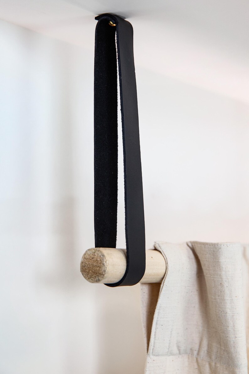 May include: A black leather strap with a wooden dowel, designed for hanging a curtain or towel. The strap is attached to the ceiling with a brass rivet. The curtain is a light beige color.