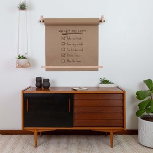 Keyaiira wall mounted kraft paper holder with veg tan leather straps and wood dowels, displaying a reusable grocery list and family command center.
