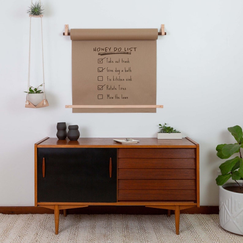 Keyaiira wall mounted kraft paper holder with veg tan leather straps and wood dowels, displaying a reusable grocery list and family command center.