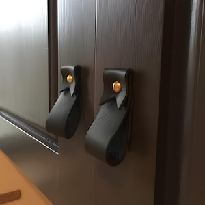 May include: Two black leather cabinet pulls with gold-colored rivets. The pulls are looped and attached to a dark brown cabinet door. The hardware provides a modern and minimalist aesthetic.