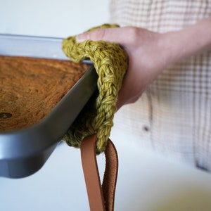 May include: A green knitted pot holder with a brown leather strap. The pot holder is being used to lift a baking pan.