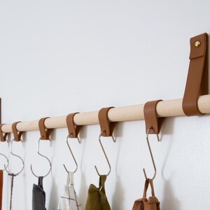 May include: A wooden rod with brown leather straps attached to the wall, with five metal hooks hanging from the straps. The hooks are holding various items, including a brown leather strap, a gray fabric item, a white and black plaid fabric item, a green fabric item, and a brown leather bag.